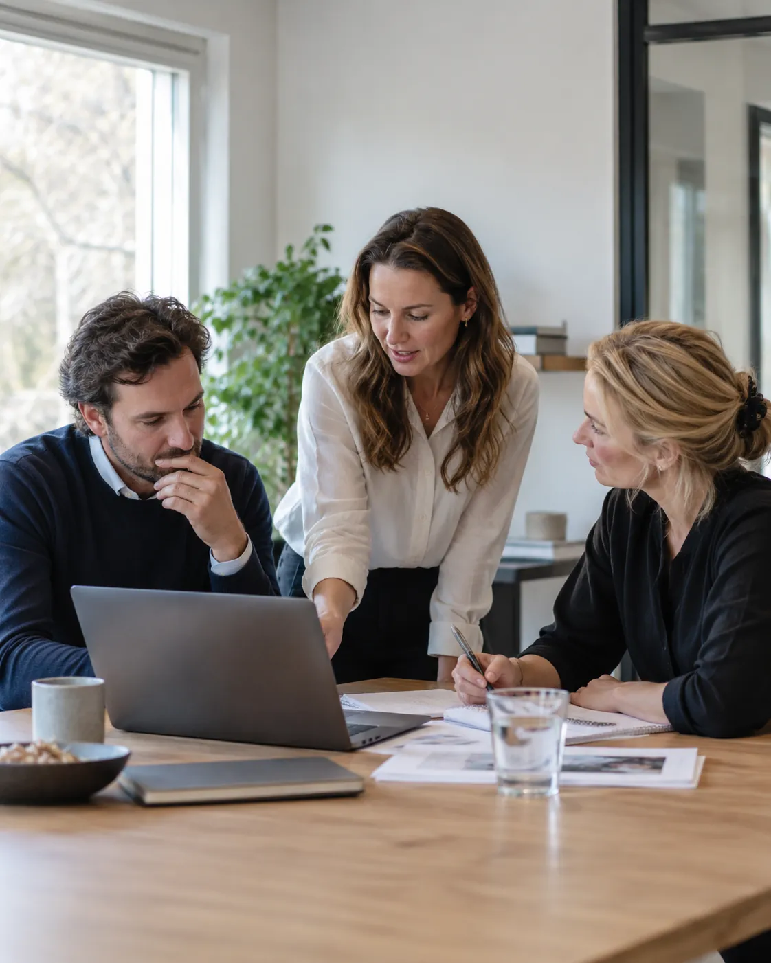 Drie collega’s bespreken documenten rond een laptop in een lichte werkruimte.